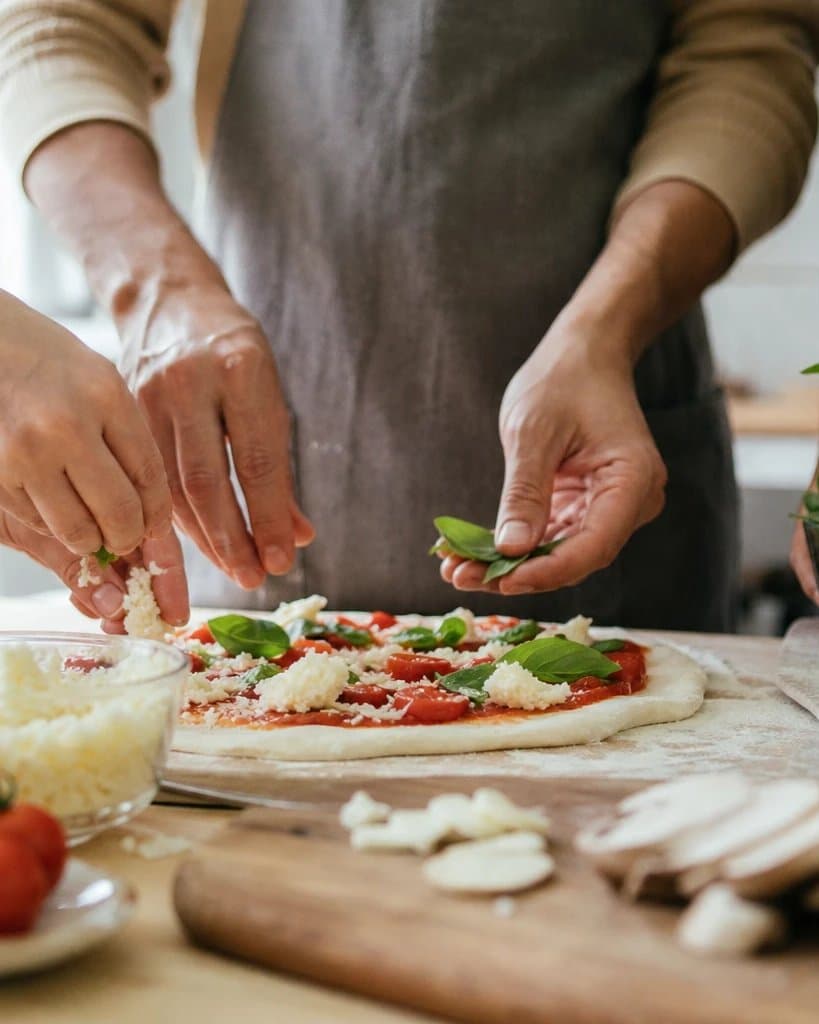 Several people making a pizza together with fresh tomatoes and basil